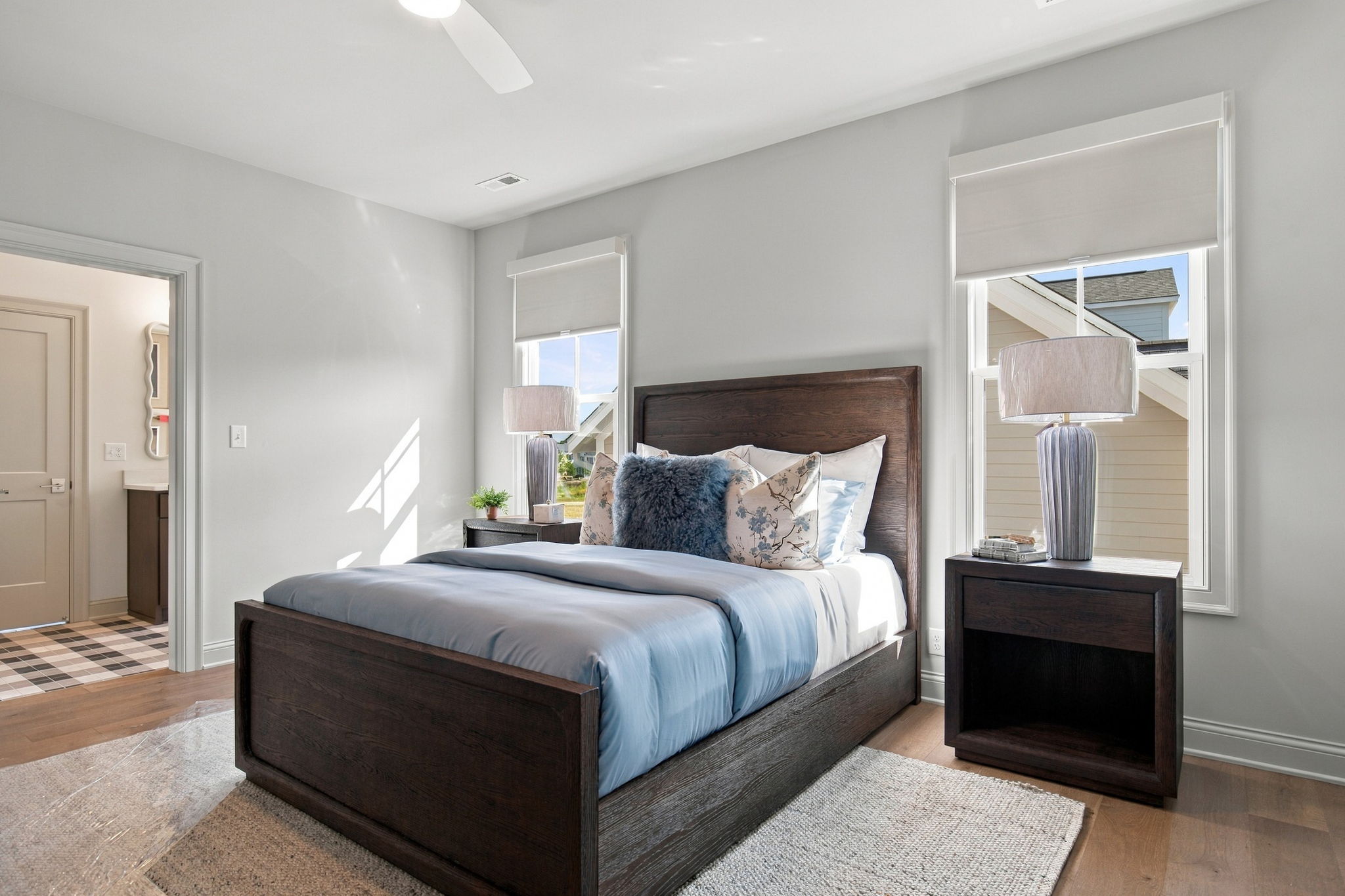 Bedroom with a dark wood bed, blue bedding, and two matching nightstands with lamps; two windows with blinds flank the bed