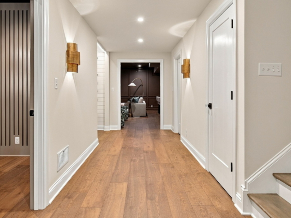 Bright hallway with wood floors and white trim, leading to a living room with a floor lamp and sofa visible at the end.