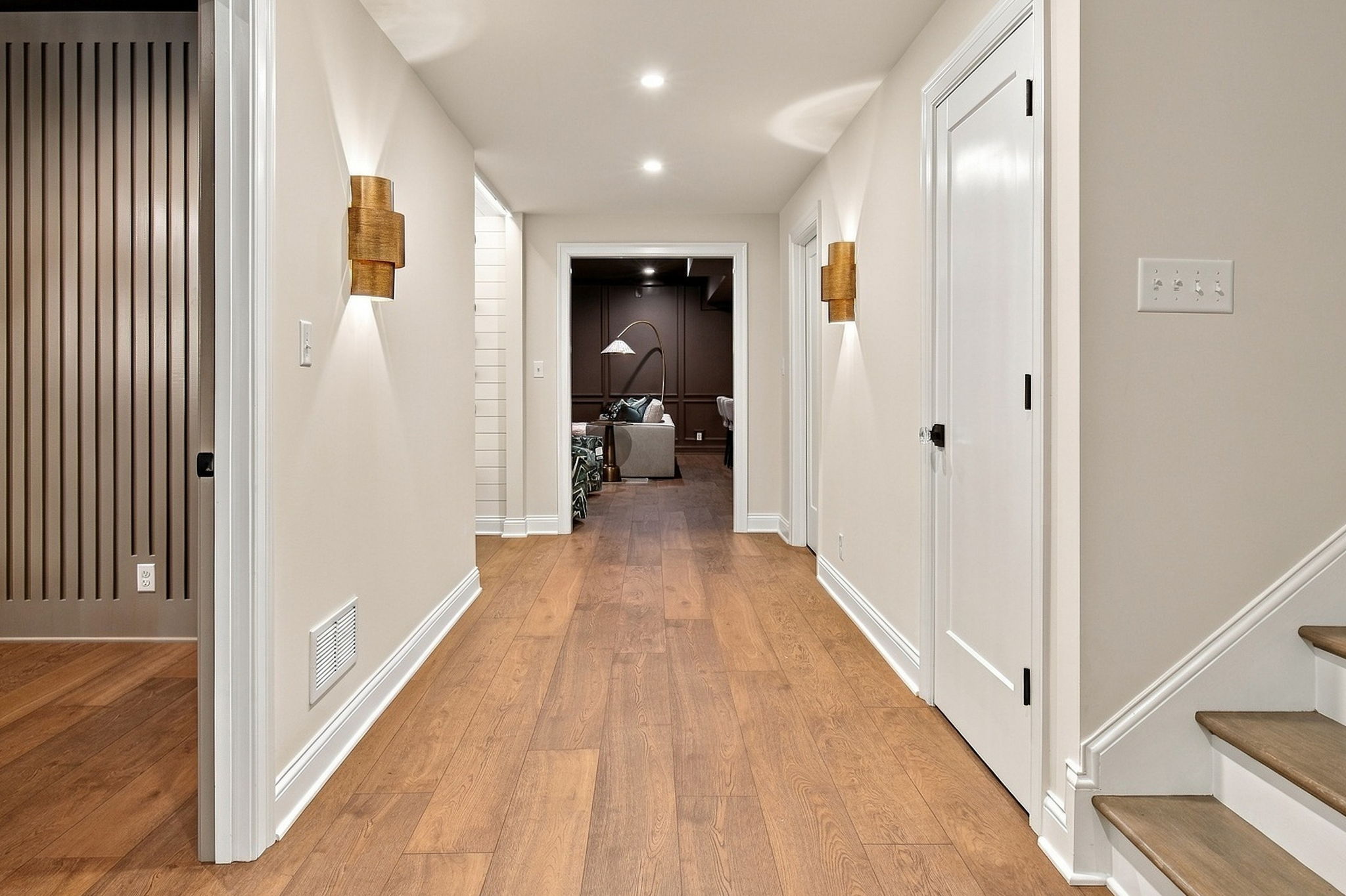 Bright hallway with wood floors and white trim, leading to a living room with a floor lamp and sofa visible at the end.