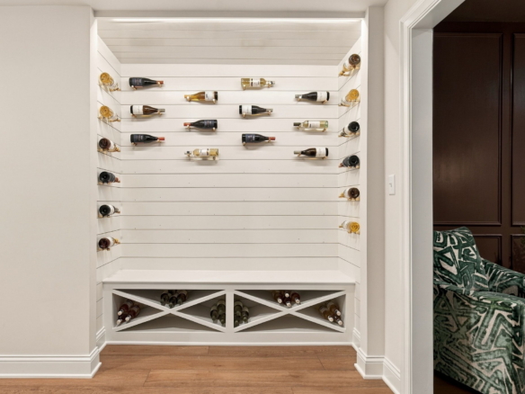 White built-in wine storage with bottles on horizontal rails and a crisscross rack below, adjacent to a living area with a green patterned sofa.