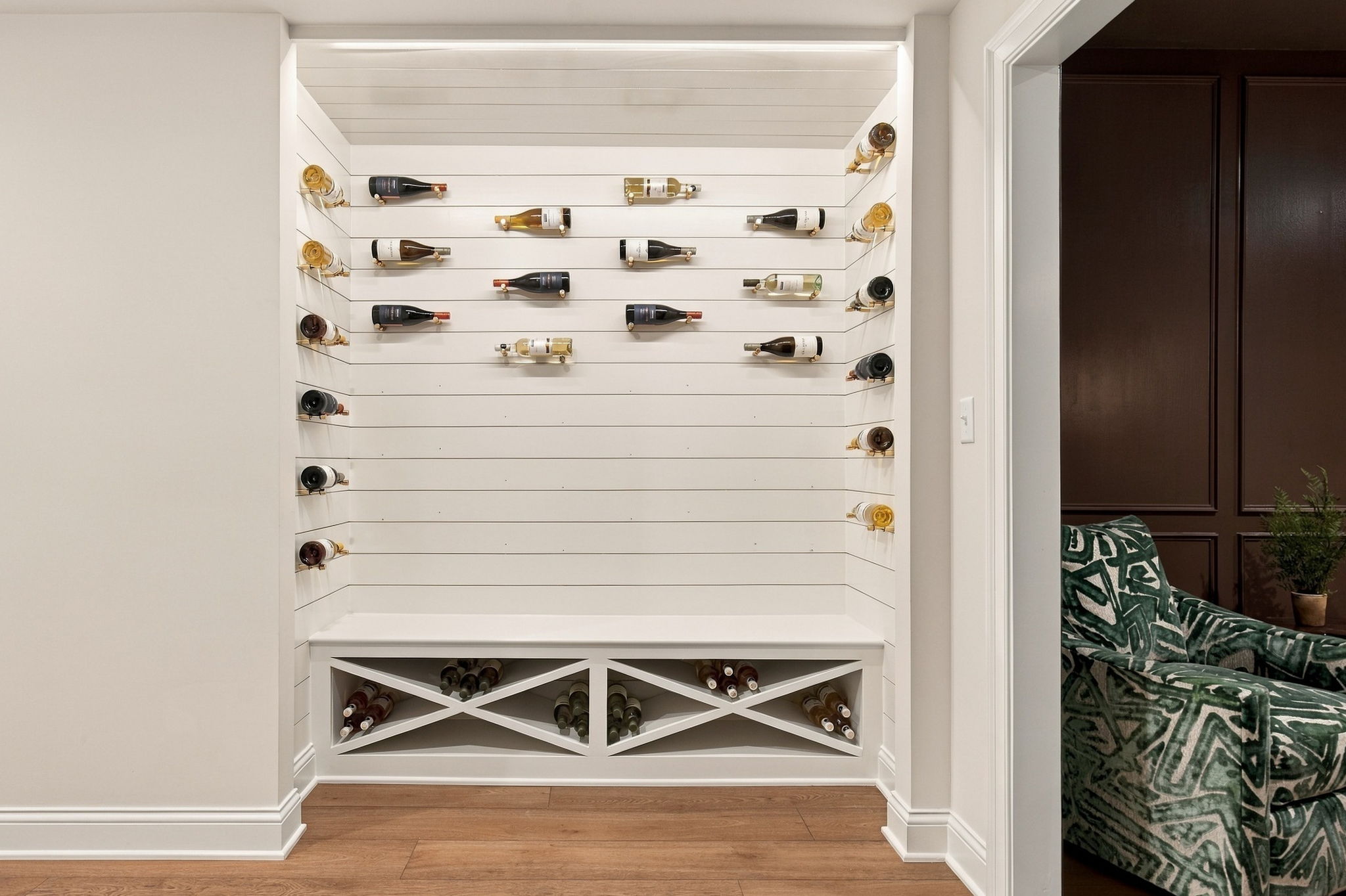 White built-in wine storage with bottles on horizontal rails and a crisscross rack below, adjacent to a living area with a green patterned sofa.