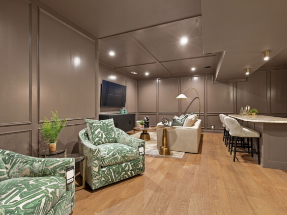 Living room with taupe paneled walls, green leaf-pattern chairs, and a TV mounted on the wall across from a beige sofa.