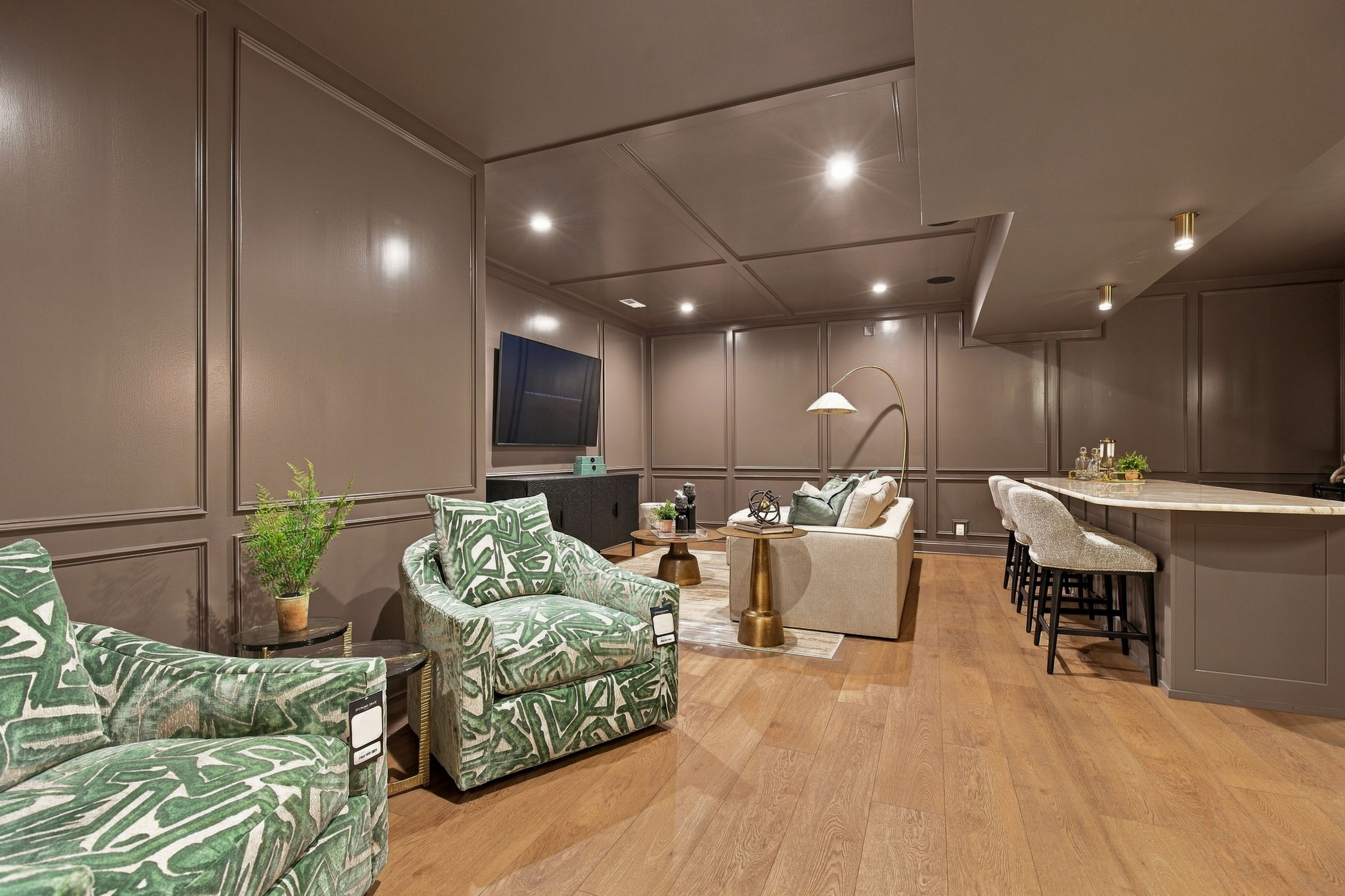 Living room with taupe paneled walls, green leaf-pattern chairs, and a TV mounted on the wall across from a beige sofa.