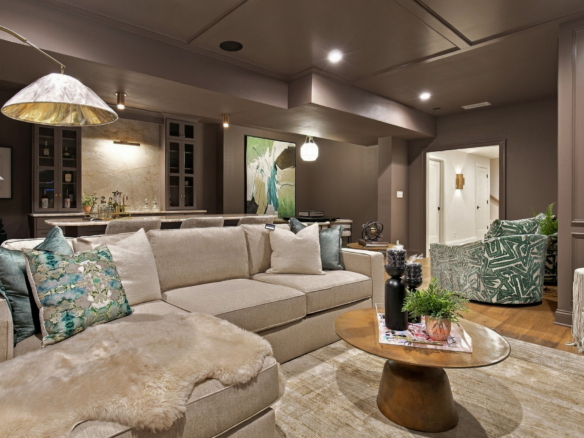 Cozy modern living room with a beige sectional, teal patterned pillows, and a fluffy throw, open to a kitchen area with pendant lights behind.