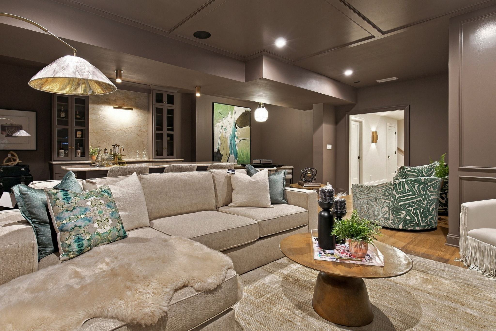 Cozy modern living room with a beige sectional, teal patterned pillows, and a fluffy throw, open to a kitchen area with pendant lights behind.