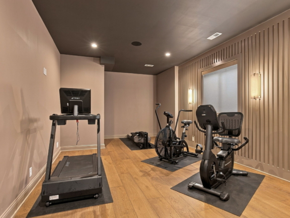 Compact home gym with two treadmills and two stationary bikes on black mats, wood floor, beige walls.