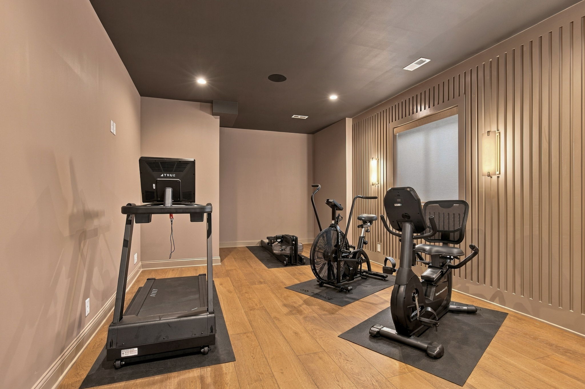 Compact home gym with two treadmills and two stationary bikes on black mats, wood floor, beige walls.