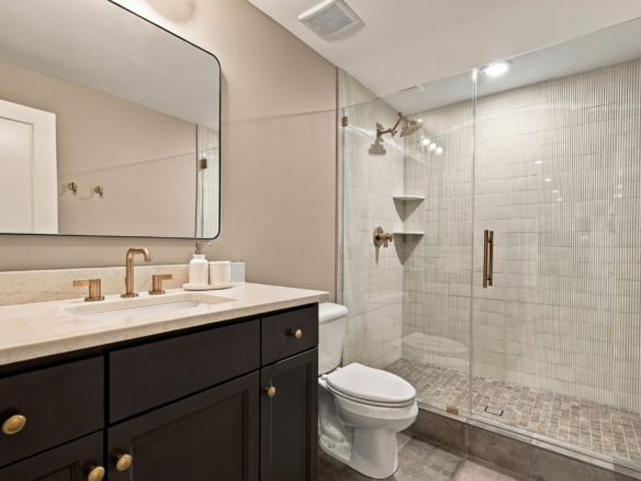 Modern bathroom with a dark wood vanity, cream countertop, and brushed brass faucet set; mirror above and a tile shower to the right.