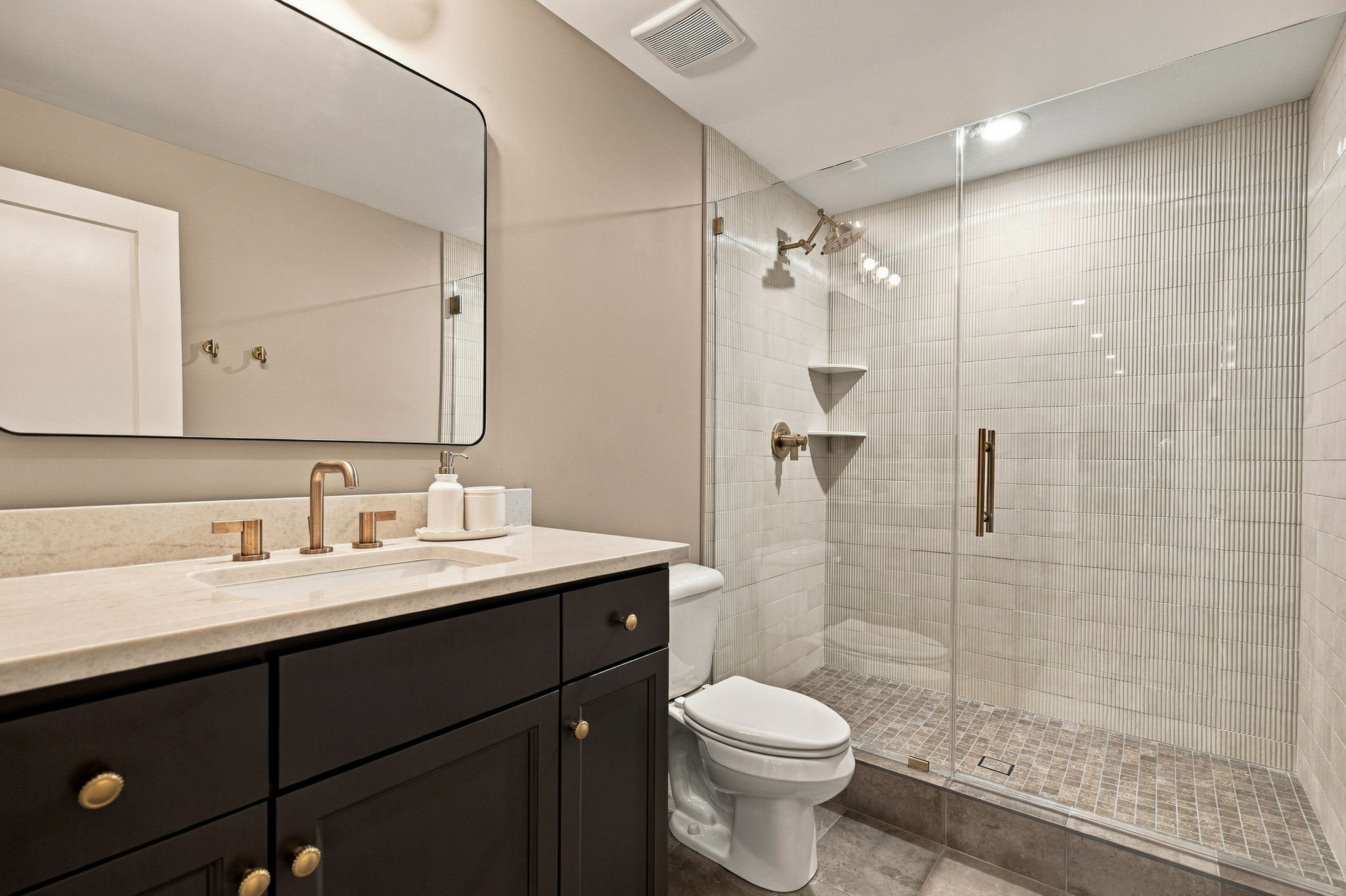 Modern bathroom with a dark wood vanity, cream countertop, and brushed brass faucet set; mirror above and a tile shower to the right.